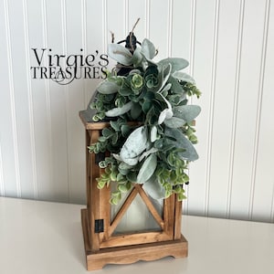 May include: A wooden lantern with a glass window and a black metal handle. The lantern is decorated with a garland of green and white foliage.