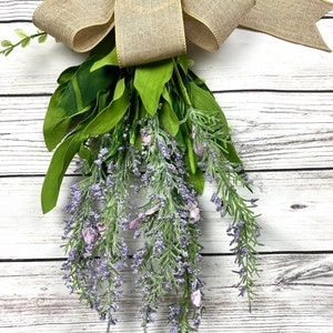May include: A bouquet of lavender sprigs with a burlap bow tied at the top. The bouquet is hanging from a white wooden surface.