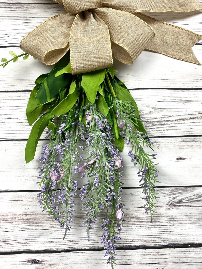 May include: A bouquet of lavender sprigs with a burlap bow tied at the top. The bouquet is hanging from a white wooden surface.