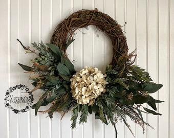 May include: A grapevine wreath decorated with faux greenery and white hydrangea blooms.