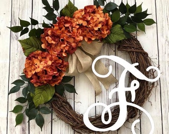 May include: A brown twig wreath with a burlap bow and orange hydrangea flowers. The wreath has a white wooden letter "F" attached to it.