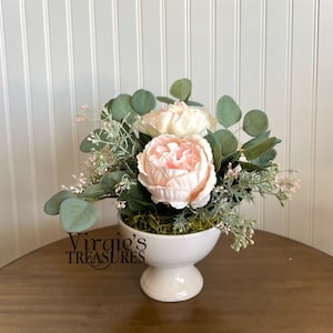 May include: A white ceramic pedestal vase with a pink and white floral arrangement. The arrangement includes two large pink peonies and eucalyptus leaves.