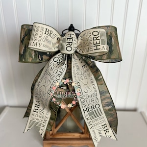 May include: A wooden lantern with a camouflage ribbon bow. The ribbon has the words "Hero", "Army", "USA", "Freedom", "Duty", "Strong", "Brave", "Service", "Thank You", and "Proud" printed on it.