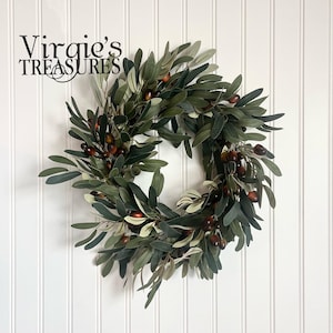 May include: A green olive branch wreath with brown olives. The wreath is hanging on a white wall with vertical wood paneling. The text "Virgie's Treasures" is in the top left corner of the image.