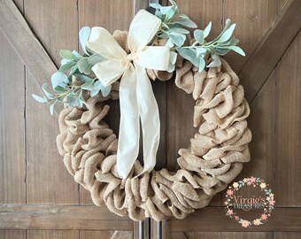 May include: A burlap wreath with a white satin bow and sprigs of eucalyptus leaves. The wreath is hanging on a wooden door.