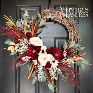 May include: A burgundy, white, and green floral wreath with a grapevine base. The wreath is decorated with burgundy flowers, white roses, and eucalyptus leaves. The wreath is hanging on a black door with the text "Virgie's Treasures" in white.