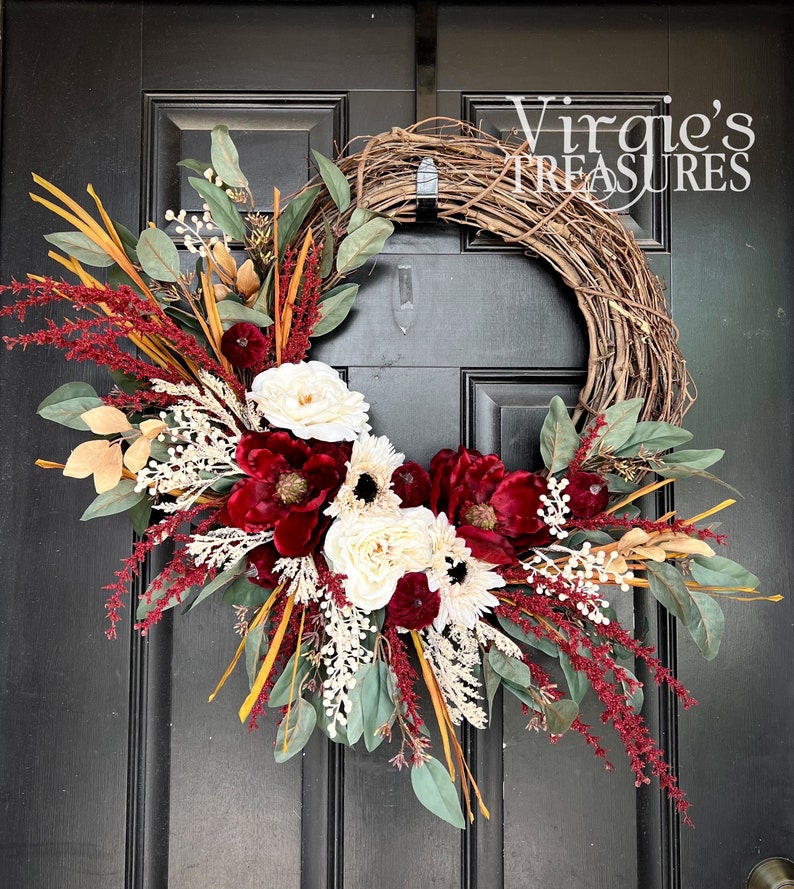 May include: A burgundy, white, and green floral wreath with a grapevine base. The wreath is decorated with burgundy flowers, white roses, and eucalyptus leaves. The wreath is hanging on a black door with the text "Virgie's Treasures" in white.
