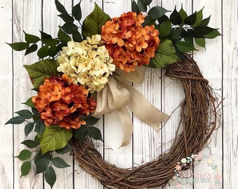 May include: A brown grapevine wreath decorated with three bunches of faux hydrangeas. The hydrangeas are in shades of cream and orange. A burlap bow is tied around the base of the flowers.