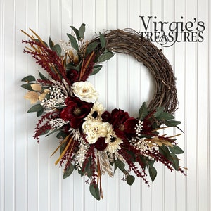 May include: A burgundy and cream floral wreath with greenery and dried grasses on a white background. The wreath is made of grapevine and is decorated with burgundy roses, cream roses, and white flowers. The wreath is accented with dried grasses and greenery.
