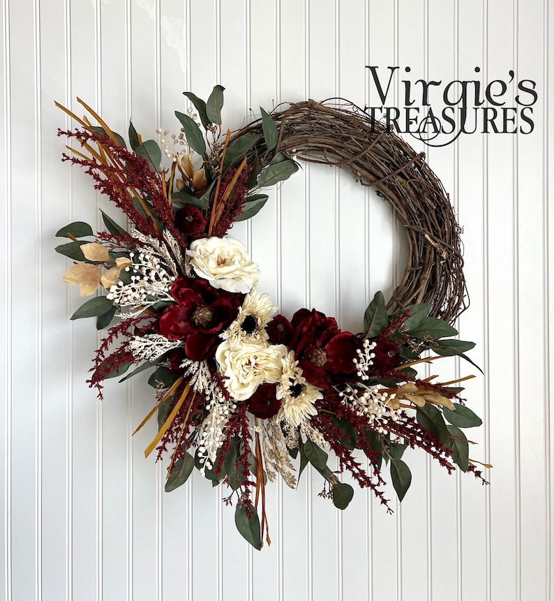 May include: A burgundy and cream floral wreath with greenery and dried grasses on a white background. The wreath is made of grapevine and is decorated with burgundy roses, cream roses, and white flowers. The wreath is accented with dried grasses and greenery.