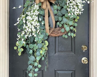 May include: A decorative wreath with a brown bow and the text "Virgie's Treasures" on a black door. The wreath features green eucalyptus leaves and white flowers, creating a natural and inviting aesthetic.