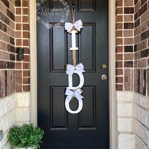 May include: A white wooden "I DO" sign with white bows hanging on a black door with a welcome mat.