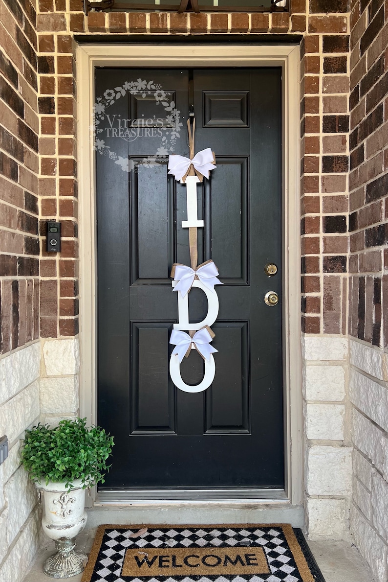 May include: A white wooden "I DO" sign with white bows hanging on a black door with a welcome mat.