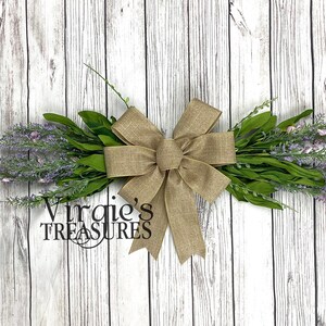 May include: A lavender wreath with a burlap bow. The wreath is tied with a brown burlap ribbon and has a sign that says "Virgie's Treasures".
