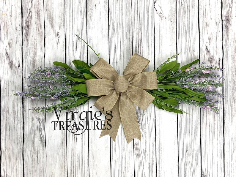 May include: A lavender wreath with a burlap bow. The wreath is tied with a brown burlap ribbon and has a sign that says "Virgie's Treasures".
