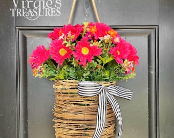 May include: A hanging floral basket filled with vibrant pink flowers with yellow centers and greenery. The basket is made of woven brown twigs and adorned with a black and white striped bow. The basket hangs from a jute rope.
