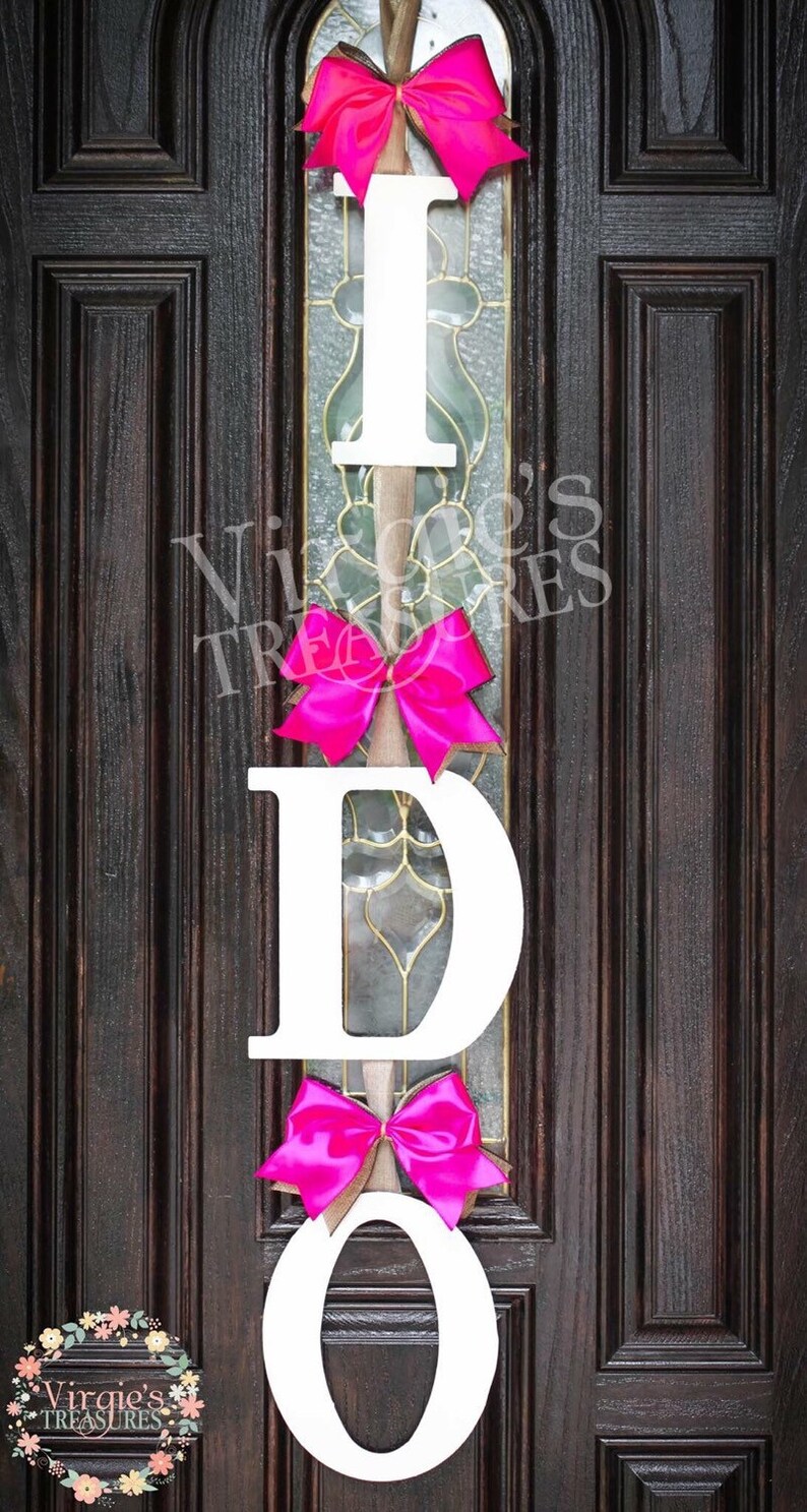 May include: White wooden letters spelling "I DO" with pink bows hanging on a dark brown wooden door.