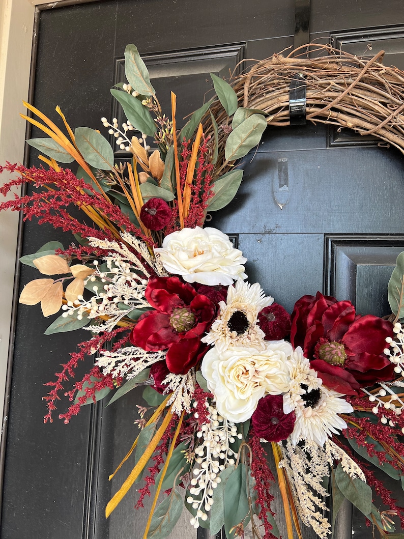May include: A burgundy and cream floral wreath with a grapevine base. The wreath features burgundy velvet flowers, cream roses, and eucalyptus leaves.