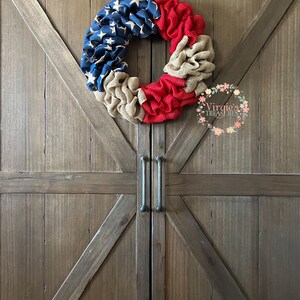 Puede incluir: Una corona de arpillera roja, blanca y azul con un patr&oacute;n de estrellas en la secci&oacute;n azul. La corona est&aacute; colgada en una puerta de madera con un acabado r&uacute;stico.