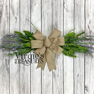 May include: A rustic wreath with a burlap bow and sprigs of lavender flowers. The wreath is tied with a large burlap bow and has a sign that says "Virgie's Treasures".
