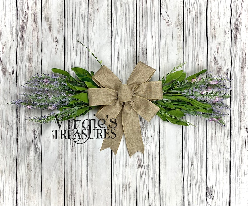 May include: A rustic wreath with a burlap bow and sprigs of lavender flowers. The wreath is tied with a large burlap bow and has a sign that says "Virgie's Treasures".