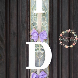 May include: A white wooden "I DO" sign with purple bows hanging on a dark brown wooden door. The sign is decorated with burlap ribbon.