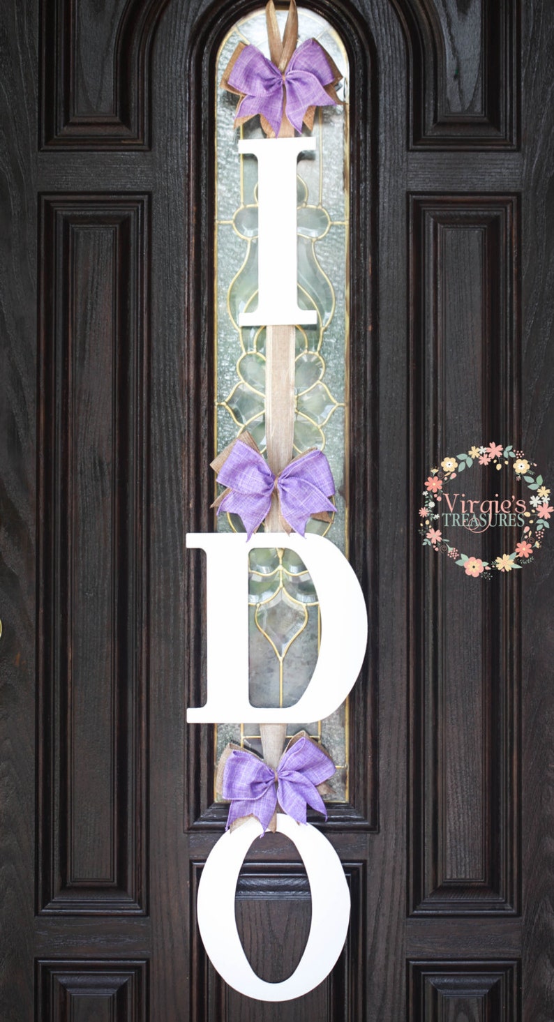 May include: A white wooden "I DO" sign with purple bows hanging on a dark brown wooden door. The sign is decorated with burlap ribbon.