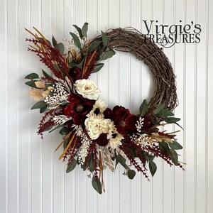 May include: A grapevine wreath decorated with burgundy and cream flowers, greenery, and dried grasses. The wreath is accented with gold accents and has a rustic, farmhouse feel.