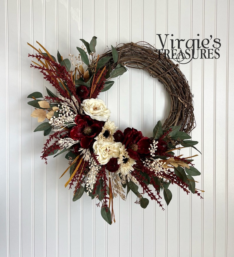 May include: A grapevine wreath decorated with burgundy and cream flowers, greenery, and dried grasses. The wreath is accented with gold accents and has a rustic, farmhouse feel.