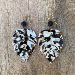 May include: A pair of earrings featuring a brown and white patterned leaf design with black lava rock beads. The earrings are made of resin and have gold-toned hooks.