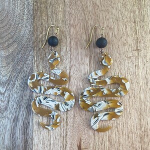 May include: A pair of gold-toned earrings with a black bead at the top and a snake-shaped design with a white and gold marbled pattern.