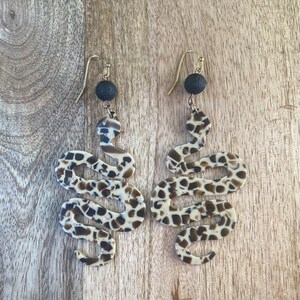 May include: A pair of dangle earrings featuring a snake design with a brown and white leopard print pattern. The earrings have black lava rock beads at the top.