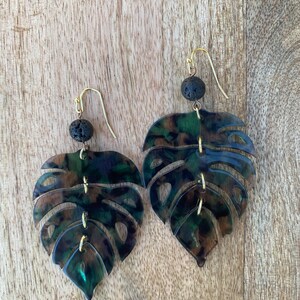 May include: A pair of earrings featuring a dark green, brown, and black monstera leaf design with gold accents and black lava rock beads.