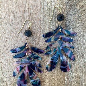 May include: A pair of dangle earrings with a layered leaf design. The leaves are made of a dark blue, pink, and white patterned resin. Each earring has a small black lava rock bead at the top.