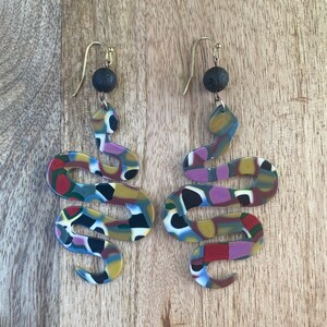 May include: A pair of colorful snake-shaped earrings with black lava rock beads. The earrings are made of polymer clay and have a gold-tone hook.