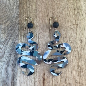 May include: A pair of dangle earrings featuring a snake design with a black, white, and brown camouflage pattern. The earrings have a black lava rock bead at the top of the dangle.