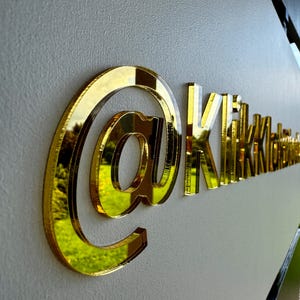 Pode incluir: Um sinal tridimensional com um grande símbolo de arroba refletor de cor dourada, seguido pelo texto Kikki com um acabamento dourado correspondente. As letras e o símbolo estão montados numa parede clara, criando sombra e profundidade.