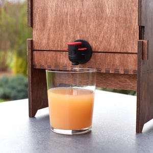 Puede incluir: Un dispensador de madera con un vaso transparente lleno de zumo de naranja. El dispensador tiene un grifo negro y rojo.