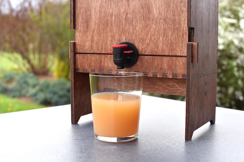 Puede incluir: Un dispensador de madera con un vaso transparente lleno de zumo de naranja. El dispensador tiene un grifo negro y rojo.