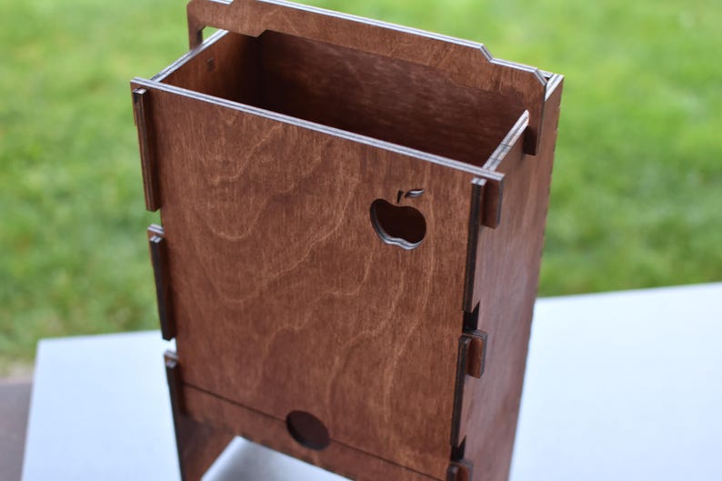 Puede incluir: Una caja de madera con un recorte de una manzana en la parte delantera. La caja tiene una tapa con bisagras y est&aacute; hecha de madera marr&oacute;n claro.