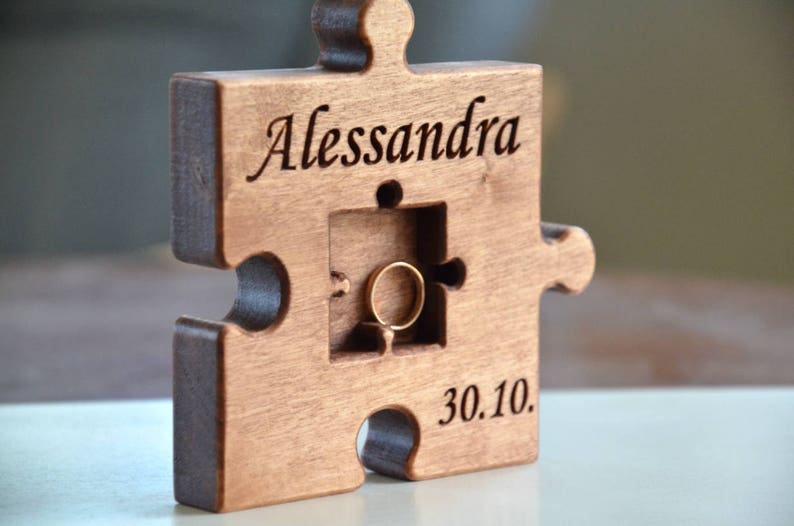 May include: A wooden puzzle piece shaped ring box with the name "Alessandra" and the date "30.10." engraved on it. The box has a cutout for a ring.