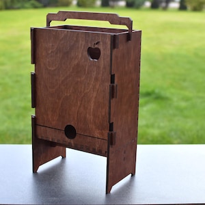 Puede incluir: Una caja de madera con un asa y un recorte de una manzana en la parte delantera. La caja es marr&oacute;n y tiene una peque&ntilde;a abertura en la parte inferior. Est&aacute; sentada sobre una superficie gris.