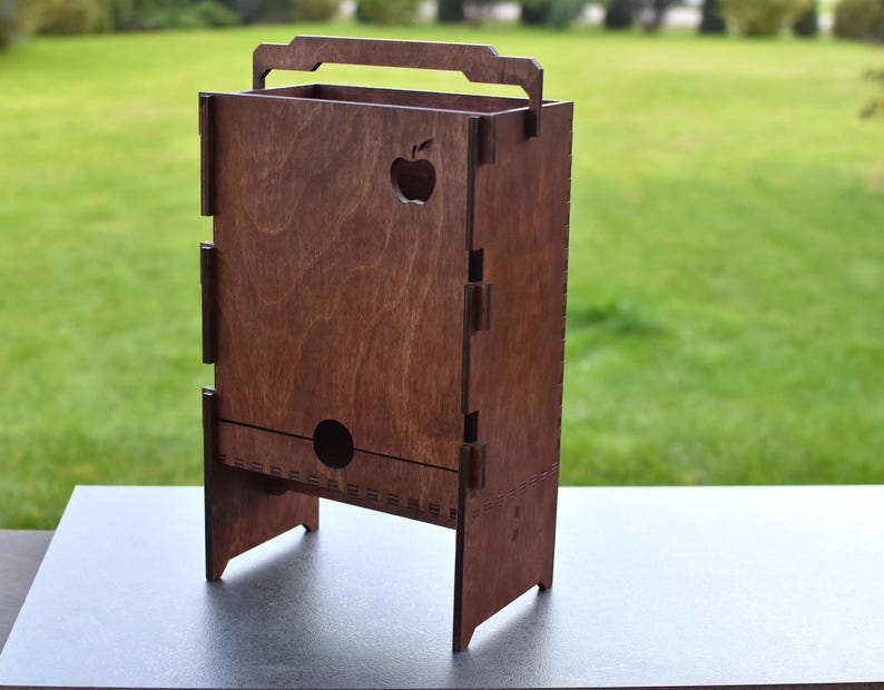 Puede incluir: Una caja de madera con un asa y un recorte de una manzana en la parte delantera. La caja es marr&oacute;n y tiene una peque&ntilde;a abertura en la parte inferior. Est&aacute; sentada sobre una superficie gris.