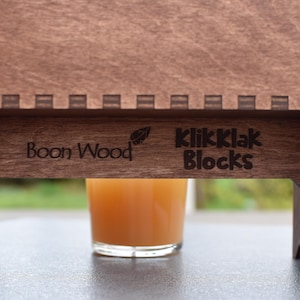 Puede incluir: Un soporte de madera con las palabras "Boon Wood" y "KlikKlak Blocks" grabadas en &eacute;l. Un vaso de zumo de naranja se encuentra sobre el soporte.