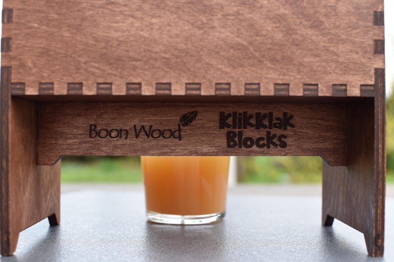 Puede incluir: Un soporte de madera con las palabras "Boon Wood" y "KlikKlak Blocks" grabadas en &eacute;l. Un vaso de zumo de naranja se encuentra sobre el soporte.