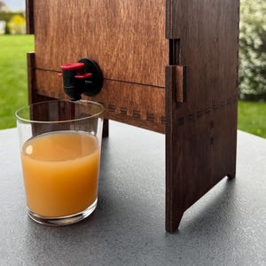 Puede incluir: Un dispensador de madera con un grifo rojo y negro, vertiendo jugo en un vaso transparente. El dispensador tiene un acabado de madera oscura y un dise&ntilde;o decorativo.
