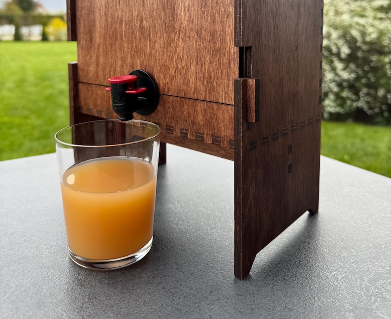 Puede incluir: Un dispensador de madera con un grifo rojo y negro, vertiendo jugo en un vaso transparente. El dispensador tiene un acabado de madera oscura y un dise&ntilde;o decorativo.