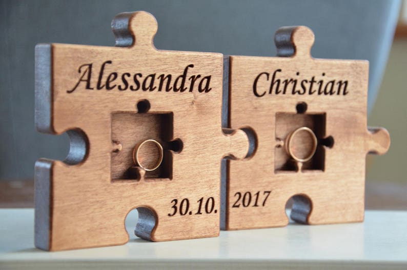 May include: A wooden puzzle ring holder with two pieces, one with the name "Alessandra" and the other with the name "Christian". The date "30.10. 2017" is engraved on the bottom of the holder.