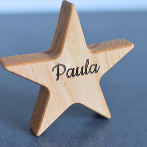 Può includere: Decorazione a forma di stella in legno chiaro con il nome "Paula" inciso in corsivo. La stella è in piedi su una superficie.