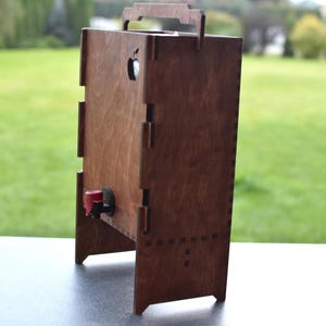 Puede incluir: Una caja de madera con un grifo rojo, dise&ntilde;ada para contener y dispensar l&iacute;quidos. La caja tiene un asa en la parte superior y un recorte en la parte delantera para el grifo. La caja est&aacute; hecha de madera oscura y tiene un acabado de madera natural.